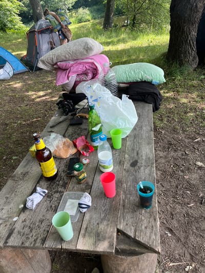 This image depicts a campsite scene, likely in Вяззеўскі сельскі Савет, Belarus. The foreground is dominated by a wooden picnic table laden with various objects, suggesting recent or ongoing activity. Among the items are several plastic cups in green and pink, empty plastic bottles, a glass bottle of what appears to be beer with a yellow label, a white plastic bag, some food packaging, two pairs of socks, a small black electronic device, and a camera. A black garment is also visible on the table.
In the background, a tent is set up, and two people appear to be resting or sleeping inside. One person is visible with their head on a pillow, and another is partially obscured. Pillows and blankets, one in pink and another light green, are arranged on or near the tent. A large tree trunk is prominent on the left side of the image. The ground is covered with dirt and grass, with fallen leaves and twigs. The overall impression is of a casual, perhaps slightly messy, outdoor gathering or camping trip. The lighting suggests it is daytime, with sunlight filtering through the trees, creating dappled shadows. The environment appears to be natural, with greenery and trees indicating a rural or park-like setting.
There is some visible text on the beer bottle label, which appears to be in Cyrillic script. Text is also visible on a small green container, possibly a spice jar, which reads "JACOBS".