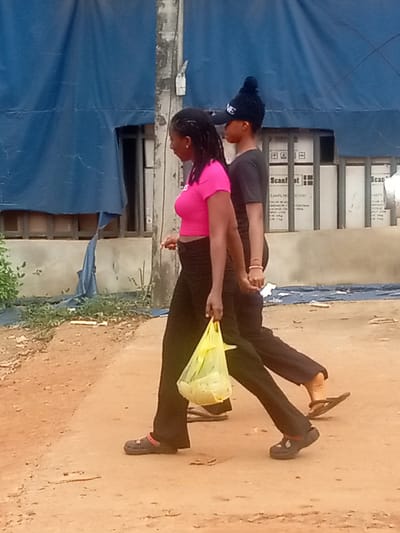 Two individuals are walking side-by-side on a dirt road in Umuokpu, Nigeria. The person on the left is wearing a pink shirt and black pants, carrying a yellow plastic bag. The person on the right is wearing a black shirt, black pants, and a dark-colored cap. Both are wearing dark-colored footwear. A blue tarp is draped over a building behind them. Boxes are stacked against the wall of the building with the tarp on it. The ground is uneven and dusty.