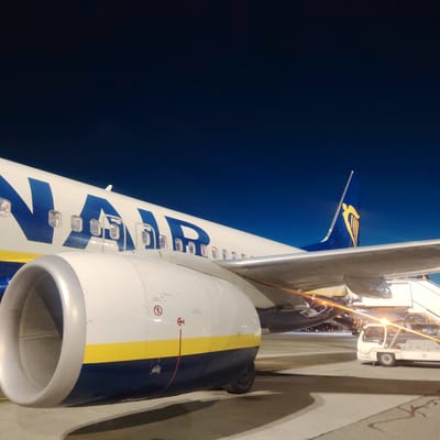 A Ryanair aircraft is depicted on an airport tarmac at night, situated in Skulte, Latvia. The aircraft, predominantly white, features a blue "RYANAIR" livery along its fuselage, partially visible on the left side of the frame, above a prominent white engine nacelle with a metallic grey intake. A horizontal yellow stripe runs along the lower part of the fuselage and engine nacelle, which also displays several warning labels and symbols. The blue vertical stabilizer (tail fin) with the distinct yellow Ryanair harp logo is visible on the right side of the frame, beyond the wing. The aircraft's left wing, silver-colored, extends into the foreground, with its underside reflecting orange artificial light.

Below the wing and near the rear of the aircraft, a white mobile passenger staircase is positioned against the fuselage, leading to an entry point. Adjacent to the staircase and beneath the wing, a white ground support vehicle with wheels is present, illuminated by a bright orange light source, creating a strong glow and reflections. The tarmac is concrete, showing shadows cast by the aircraft and ground equipment. The sky is dark blue, indicative of nighttime conditions, with distant, blurred lights visible in the background. No individuals are explicitly visible in the image.