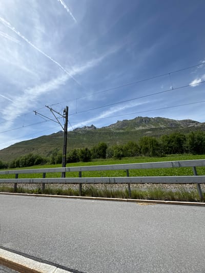 The image shows a view from a road in Andermatt, Switzerland, looking towards a green mountain range under a blue sky with wispy white clouds and contrails. A grey asphalt road is in the foreground, bordered by a metallic guardrail. Beyond the guardrail, a green grassy field stretches towards a line of lush green trees, which form the base of the mountain. A single utility pole with overhead electrical wires for a train is visible in the mid-ground. The mountain itself is covered in green vegetation with rocky outcrops visible at its peak. The scene appears to be a scenic landscape captured during daytime.