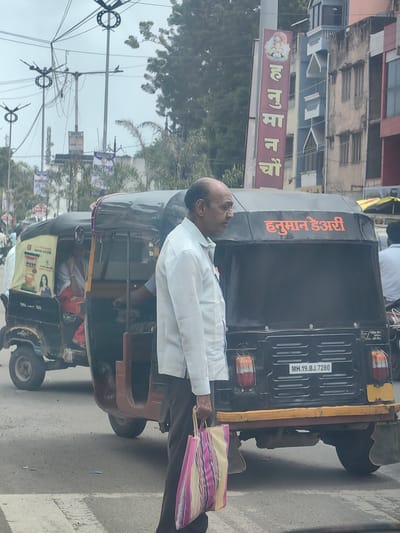 This is a street-level, medium-shot photograph taken from a vehicle, capturing a street scene in Datala, India. In the foreground, a bald man in a white shirt and dark pants walks towards the right, carrying a striped tote bag. To his right, a black auto-rickshaw with a yellow stripe across its rear and orange-red lettering is parked, bearing the license plate "MH.19.BJ.7280". The lettering on the auto-rickshaw reads "हनुमान डेअरी" (Hanuman Dairy). Behind the auto-rickshaw, another auto-rickshaw is partially visible, with a woman inside. In the background, buildings with signs are visible, including a vertical red sign with a deity image and text "हनुमान" (Hanuman) in yellow. Utility poles and electrical wires crisscross the sky, which is overcast. The scene depicts a typical urban environment with vehicular traffic and commercial signage.