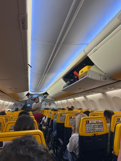 The image depicts the interior of an airplane cabin, viewed from a passenger's perspective looking forward. The seats are upholstered in yellow and dark blue. Overhead compartments are open, revealing luggage, including a yellow suitcase and red items. A man wearing a light brown t-shirt and red shorts is standing in the aisle, gesturing with his right hand. Several passengers are seated, facing forward, with their heads and shoulders visible. The cabin is illuminated by blue ambient lighting from the ceiling. Safety information cards are visible on the back of the yellow seatbacks. The context provided suggests this scene is at Orio al Serio, Italy, indicating it is likely the interior of a commercial airliner at or near that location.