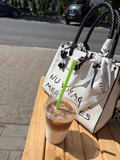 In Pastavy, Belarus, on a sunny day, an iced coffee in a plastic cup with a green straw rests on a wooden bench. Behind the coffee, a white handbag with black braided handles and a black and white patterned scarf tied to it sits on the same bench. The handbag has black text on it, some of which is partially visible. In the background, a paved sidewalk is visible, beyond which is a street with a few cars and trees lining the other side. The scene suggests a moment of relaxation outdoors.