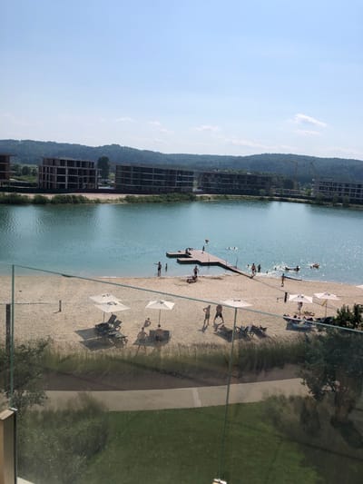 The image shows a man-made lake with a sandy beach, located in the city of Pidhirtsi, Ukraine. The lake is bordered by trees and hills in the background, with several unfinished multi-story buildings visible on the left side of the image. On the beach, there are several umbrellas and lounge chairs, suggesting a recreational area. People are seen swimming and sunbathing on the beach and in the water. A wooden pier extends into the lake, and some individuals are standing on it. The overall scene depicts a warm, sunny day at a lakeside resort or development.