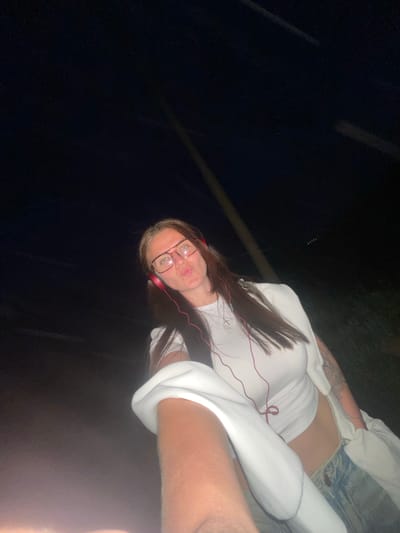 A young woman is posing for a photo outdoors at night. She is wearing glasses, headphones, a white t-shirt, and light-wash denim shorts. She has long, dark hair with blonde streaks in the front. Part of another person's arm is visible on the right side of the frame. The location is assumed to be in the city of Molokovo, Russia.