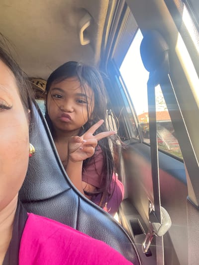A young girl in a pink shirt makes a peace sign with her fingers as she sits in the backseat of a car. She has dark hair and is pouting her lips. To her left, partially visible, is the face of an adult woman in the driver's seat, wearing a bright pink garment. The car interior is visible, including black leather seats and a beige headliner. Sunlight streams in through the window on the right side of the frame. The background visible through the car window shows a blurred outdoor scene with houses and vegetation. The image appears to be taken in Gerokgak, Indonesia, based on the context provided.