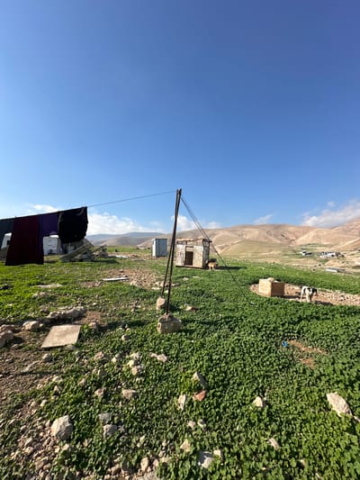 The image depicts an expansive, rural landscape in the Arvot Hayarden Regional Council, Palestinian Territory, under a clear blue sky with a few distant white clouds. The foreground consists of uneven, rocky terrain interspersed with dense patches of bright green, low-lying vegetation. A light-colored square slab of concrete is visible on the ground in the lower-left quadrant.
In the mid-ground, a clothesline stretches diagonally from the left edge of the frame towards a vertical pole anchored by two concrete blocks. Several pieces of dark-colored laundry, including maroon, purple, and black garments, are hanging to dry on the line. To the left of the pole, a white, possibly tent-like, temporary structure is visible. Further right, a small, silver-colored metal shed or portable toilet stands beside a more substantial, rectangular, rudimentary structure constructed from various materials, featuring an open doorway and a partially covered flat roof. A tan-colored dog with dark markings is standing near the entrance of this rudimentary structure.
Towards the right mid-ground, a black and white cow is visible amidst the vibrant green vegetation, near a small, square, concrete-like box structure. The background features rolling, arid, light-brown hills and mountains stretching across the horizon, with sporadic greenery on their lower slopes and faint indications of distant structures or settlements nestled within the landscape. No people are present in the scene.