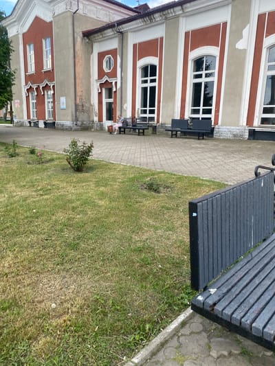 This image depicts an outdoor scene in Stadnia, Ukraine, featuring a building with a red and beige facade and white trim. Several windows with white frames are visible, some arched. In front of the building, there is a paved walkway and a grassy area. A dark wooden bench is positioned on the walkway, and another similar bench is situated closer to the building, near a window. A young person wearing a pink outfit is seated on the second bench, appearing to be engaged in an activity. The lighting suggests it is daytime. The overall atmosphere appears calm and ordinary, with no indication of a specific event.