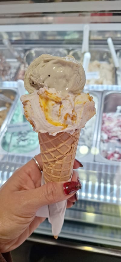 The image shows a person holding an ice cream cone with two scoops of gelato in front of a gelato display case. The first scoop on the left is a light green color, while the second scoop has a white base with swirls of yellow and orange. The ice cream cone is a traditional waffle cone. The person's hand is visible, with a ring on one finger and polished fingernails. The background shows a gelato display case with various flavors of gelato visible in metal containers. The reflection on the glass suggests that the location is indoors, likely an ice cream shop. While the image itself does not explicitly pinpoint the location as Santa Cruz de Tenerife, Spain, this information could be derived from the context in which the image was provided.