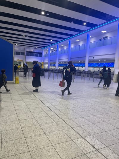 The image depicts an indoor scene within what appears to be a modern airport terminal, characterized by its spaciousness and contemporary design. The ceiling features a distinctive pattern of alternating white and black panels, accented by a bright blue LED light strip that runs diagonally across the upper portion of the frame. Numerous recessed square lights illuminate the area, and robust white pillars support the structure. The floor is covered in light grey speckled tiles, arranged in a grid pattern.
Several individuals are present, engaged in typical airport activities. In the mid-ground, a person wearing dark clothing, including a jacket and trousers, walks away from the camera, carrying a dark backpack and a red gift bag in their left hand. Text, possibly "IRON MAN," is visible on their backpack. Further back, two more people in dark attire with backpacks walk in the same direction. Closer to the foreground, another individual stands, looking down, potentially at a mobile phone. This person wears a long dark coat or skirt, a dark top, and carries a dark backpack, holding a red item in their hand. In the far foreground, partially obscured, another person walks towards the right, also wearing dark clothing and a backpack, with athletic shoes featuring white stripes. In the background, rows of check-in counters with blue-lit screens and queue barriers are visible, where more people are gathered or passing through.
Notable details include a yellow caution sign, likely indicating a "wet floor," placed on the tiled surface in the mid-foreground. On one of the digital screens above the check-in counters, the time "16:48" is clearly displayed, suggesting late afternoon. The overall atmosphere appears functional and busy, consistent with an airport environment near Horley, United Kingdom.