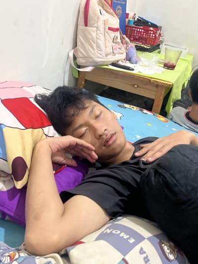 In a bedroom, a young man with short black hair sleeps on his side, facing the camera. He is wearing a black t-shirt and black pants, and his left arm is bent, with his hand resting on his chest. He is on a bed with colorful patterned sheets. The bed is positioned against a white wall. To the right of the bed, a wooden table is visible, covered with a lime green cloth. On the table are various items, including a pink backpack, a red basket filled with stationery, a purple toy, and a glass with a reddish liquid. Another person, partially visible, is seated at the far right of the frame, in the background. The scene appears to be in a residential setting.