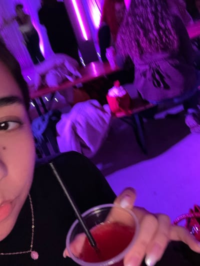 A blurry, angled selfie shows a person with fair skin, dark eyebrows, and visible eyeliner. A clear plastic cup filled with a red liquid and a black straw is held nearby. The background is dark with purple lighting. A Halloween mask is visible near the top left. A white-sleeved arm and part of a shelf appear to be in the middle.