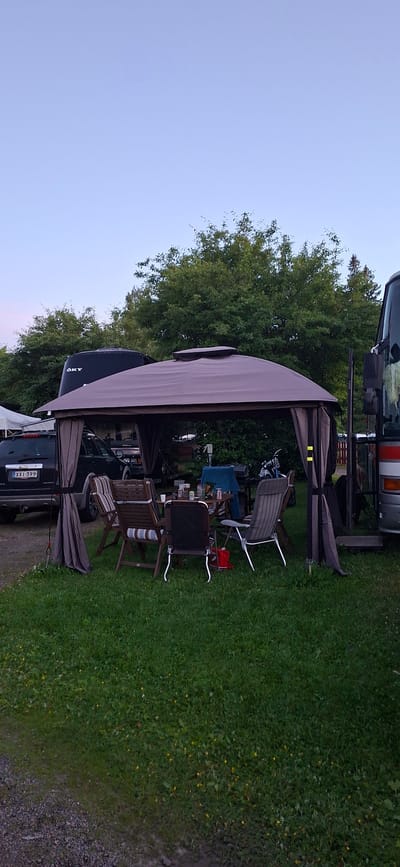 The image depicts an outdoor scene at a campsite or recreational area, possibly in Salpakangas, Finland, given the context. In the foreground, a brown canopy tent is set up, providing shelter over a picnic table laden with various items including glasses, bottles, and what appear to be condiments. Several chairs are arranged around the table, some occupied by cushions. To the left of the tent, a dark-colored SUV with license plate "XXI-399" is parked. In the background, lush green trees form a dense backdrop. A large, dark vehicle, possibly an RV or bus, is partially visible on the right side of the image, with a person visible near its entrance. The ground is covered in green grass, with a gravel path leading into the scene from the lower left. The sky is a clear, pale blue, suggesting it is either early morning or late evening.
