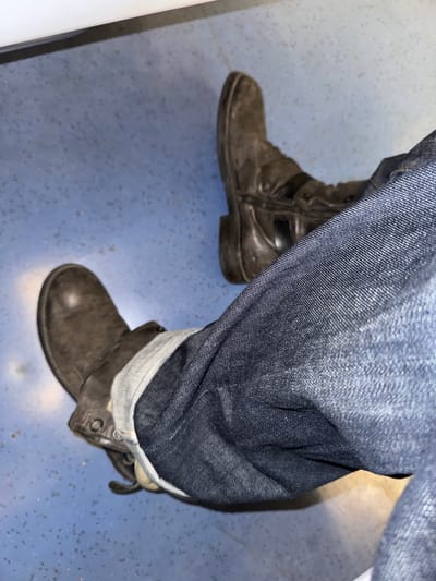 A close-up shot captures the lower half of a person wearing blue denim jeans and dark, rugged boots. The jeans are cuffed at the ankles, revealing a lighter inner fabric. The boots appear to be made of dark leather or suede, with visible wear and a sturdy construction; the boot on the left features a side zipper. The person is standing on a speckled blue-grey floor, which suggests an indoor public setting like a train, bus, or building interior. Given the context of Palaiseau, France, this floor could be found in a public transport vehicle or station. The lighting appears artificial, consistent with an indoor environment. No specific activity is discernible beyond the person standing or sitting. There is no visible text.