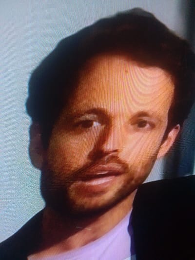 The scene is located in the city of Mista Ali, Nigeria. This image displays a close-up, head-and-shoulders portrait of an adult male, captured from a digital screen. The subject has a light-to-medium skin tone, dark hair, and a neatly trimmed beard and mustache. He is observed wearing a dark jacket or suit over a light-colored shirt, possibly lavender or pink. His eyes are open and directed forward, with his mouth slightly ajar. The background is out of focus, with lighter tones to the left and darker, indistinct areas to the right. A visible pixel grid indicates the image is a photograph of an electronic display.