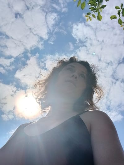 In Maslova Pristan', Russia, a low-angle, over-the-shoulder shot captures a woman looking upwards. The sun is bright and partially obscured by clouds, creating lens flare. Green leaves from a tree are visible in the upper right corner of the frame, contrasting with the blue sky and scattered white clouds. The woman has medium-length, curly brown hair. She is wearing a dark-colored top, and her face is partially in shadow due to the angle of the sun. The overall impression is one of being outdoors on a sunny day.