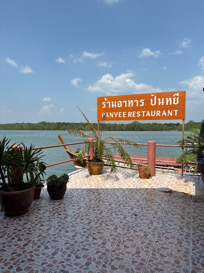 The image displays an outdoor patio area of the Panyee Restaurant in Ko Panyi, Thailand, situated above a body of water. A prominent orange wooden sign, supported by two brown posts, is centrally positioned. The sign features white Thai script "ร้านอาหาร ปันหยี" above "PANYEE RESTAURANT" in white English capital letters. The foreground consists of a tiled floor exhibiting a mosaic pattern of irregularly shaped beige, brown, and light grey pieces. Several potted plants are arranged on the patio: two large, dark brown pots with broad-leafed plants are on the left, a lighter brown pot containing a palm-like plant is directly in front of the sign, and a smaller brown pot sits to its right. A red-brown metal railing runs horizontally, separating the patio from the water. In the midground, a wooden boat with an ornate, gold-brown prow is partially visible on the left, moored at water level next to the patio structure. The water is a greenish-blue. The background reveals a dense, dark green forested landmass, likely mangroves, extending across the horizon, with distant hills visible. The sky is clear blue with sparse white cumulus clouds. No individuals or specific actions are depicted.