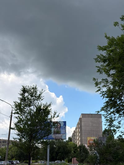 The image depicts a cloudy sky with dark gray clouds dominating the upper portion and lighter, white cumulus clouds and patches of blue sky visible lower down. A tall, multi-story, concrete apartment building with a repetitive facade is visible on the right side of the frame, partially obscured by trees. In the foreground, a large tree with dark green foliage stands to the left of the building. A billboard is positioned behind this tree, featuring a portrait of a person with glasses and text. Another smaller, circular advertisement with orange and yellow hues is affixed to the side of the apartment building. The street level below the building shows parked cars and some sparse vegetation. The overall scene suggests an urban environment in Novotroitsk, Russia, possibly on an overcast or stormy day.