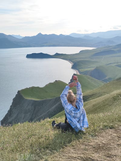 A woman with blond hair pulled back in a bun, wearing a blue and white plaid shirt and black pants, sits on a grassy hilltop with her arms raised above her head. She faces a scenic view of a bay with a long, sandy beach, surrounded by green, rolling hills and mountains in the distance. The sky is overcast. The location is in the city of Коктебельский поселковый совет, Ukraine.