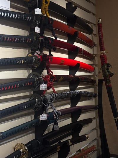 A collection of ten Japanese swords, primarily katana and wakizashi, are displayed horizontally on a light-colored wooden slatted wall within an establishment in Kanazawa, Japan. Each sword is held by black, L-shaped wall mounts. To the far right, a single Japanese sword stands vertically on a black floor stand against a textured, light brown wall.

The horizontal display features swords with varying handle (tsuka) and scabbard (saya) colors and designs. From top to bottom:
1.  A sword with a black handle featuring white accents and a dark tsuba. A white tag on its handle reads "Kondo Isami." Another tag indicates "¥32,000." Its saya is mostly obscured by the sword above, but a red saya is visible beneath it.
2.  A sword with a black handle featuring red accents and a dark, circular tsuba. Its saya is red with subtle black patterns.
3.  A sword with a black handle featuring red accents and a dark, intricately designed tsuba. Its saya is bright red with three distinct gold circular symbols. A white tag displays Japanese text and "¥24,000 (tax inc)."
4.  A sword with a black handle featuring black and red accents. Its saya exhibits a marbled pattern of dark brown and red.
5.  A sword with a black handle and a tsuba. Its saya has a dark gray and black marbled or patterned finish.
6.  A sword with a black handle. Its saya is a dark teal or greenish color. A white tag with Japanese text indicates "¥24,000."
7.  A sword with a black handle. Its saya is purple with subtle patterns.
8.  A sword with a black handle. Its saya is black.
9.  A sword with a black handle featuring gold ornamentation. Its saya is black with gold ornamentation near the tip.
10. A sword with a black handle featuring gold ornamentation. Its saya is red with gold patterns.

On the upper left of the image, a white informational sign is partially visible, stating "se Hijikata Toshizo's favorite [sword] made by Izuminokami."

The vertically standing sword on the right has a red, diamond-pattern wrapped handle with a gold pommel cap and a gold tsuba. Its scabbard is black, adorned with a green and gold braided cord (sageo). The environment is an indoor setting with warm, artificial lighting. No individuals are present.