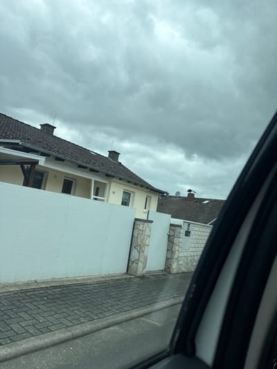 The image depicts a residential street scene under a cloudy sky in Neukirchen, Germany. In the foreground, the edge of a vehicle's interior is visible, framing the view. Beyond the vehicle, a paved sidewalk, constructed of interlocking rectangular bricks, runs along the street. Adjacent to the sidewalk is a low wall, partly constructed of white panels and partly of stacked stone blocks. Behind the wall, a pale yellow house with a dark tiled roof is visible. The house has several windows and a chimney on its roof. Further down the street, another house with a similar roof is partially visible, also featuring a chimney. The overall environment suggests a typical suburban residential area.