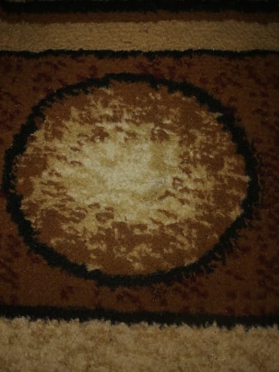 The image is a close-up, high-angle shot of a carpet with a brown, beige, and black pattern. The dominant feature is a large oval shape in the center, outlined in black and filled with a lighter brown and beige speckled pattern. Several horizontal bands of varying widths and colors are visible above and below the oval. The texture of the carpet appears to be slightly worn, with visible fibers and dust. There is no indication of the location in Juma, Uzbekistan, or any people or actions depicted. The image focuses solely on the intricate details of the carpet's design and texture.
