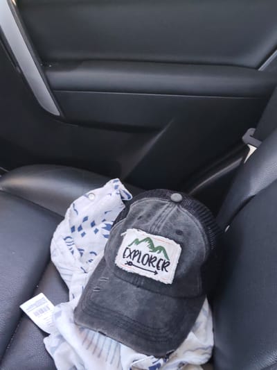 This media file shows a close-up, angled view of a dark gray, distressed trucker hat and a white and blue patterned cloth, likely a baby blanket, resting on a black leather car seat. The hat has a patch on the front with the word "EXPLORER" in green text, superimposed over a depiction of mountains and an arrow pointing right. The cloth has a geometric pattern of blue diamonds and stripes. A white tag with a barcode is attached to the cloth. The background is the interior of a car, with black leather door panels visible. The lighting suggests it is daytime, possibly in bright sunlight given the reflections on the leather. There are no people visible. The scene appears to be set inside a vehicle. The visible text includes "EXPLORER" on the hat and text on the white tag, which is too small to be legible.