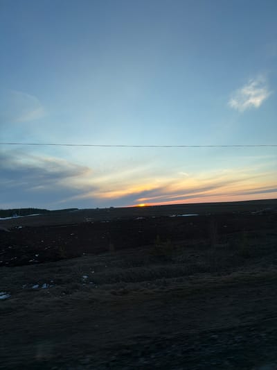The image shows a landscape in what appears to be near Дулесово, Russia based on geographic features. The sky occupies a significant portion of the image, displaying a gradient from clear blue to a sunset with orange and yellow hues. Clouds are visible in the sunset portion of the sky. A thin wire or cable stretches vertically across the left side of the image. The lower portion of the image depicts a dark, possibly barren field or land. There are some details in the field such as what seems like the beginning of trees. The scene is likely photographed at dusk or dawn.