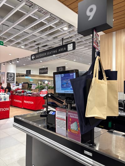 This media file captures a detailed view of a retail store's checkout and self-packing area in Kelapa Dua, Indonesia. The scene is brightly lit with artificial lighting and features a modern design.

In the foreground and midground, several red display tables are visible, each prominently labeled "PROMOTION," showcasing various products. To the left, a large, dark grey sign with a white illuminated number "9" is fixed to a pole.

Central to the image are two light-colored reusable shopping bags resting on a counter, with a dark blue bag beneath them. Behind these, a digital screen displays "Counter Closed" and an advertisement in Indonesian: "Menangkan Tiket Marathon ke Berlin!" (Win a ticket to the Berlin Marathon!).

Several signs provide information and promotions. One sign offers "GRATIS VOUCHER Rp20.000" (Free Voucher Rp20,000), while another nearby sign indicates "GRATIS VOUCHER Rp50rb" (Free Voucher Rp50,000). An important "ANNOUNCEMENT" in Indonesian, under the "AEON STORE ALAM SUTERA" banner, states that "mulai tanggal 24 Oktober 2025 layanan packing kardus tidak lagi dilakukan di area kasir. Pelanggan dapat menggunakan area self-packing yang telah kami sediakan untuk keperluan tersebut." (Starting October 24, 2025, cardboard packing services will no longer be provided at the cashier area. Customers can use the self-packing area provided for this purpose.)

Signs pointing with arrows indicate "OUTDOOR SEATING AREA" and "SELF PACKING," which features an icon of hands packing a box. Further into the store, a "BENTO" sign is visible. A few customers are present, some appearing to be at the self-packing stations or browsing promotions. The ceiling features a distinctive pattern of white horizontal slats with integrated lighting.