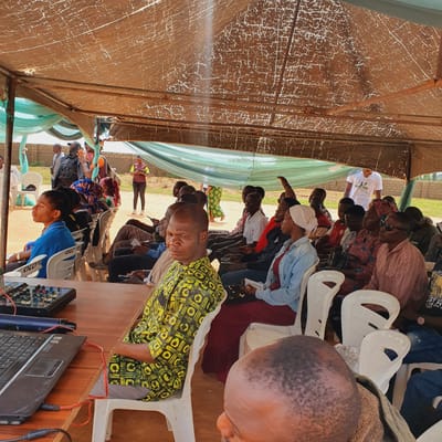 This media file depicts an outdoor gathering in Unguwar Saya, Nigeria, likely a community event or religious service taking place under a makeshift shelter. The shelter is constructed with a textured brown tarp for a roof, supported by metal poles, and draped with light green fabric on the sides.
The primary subjects are a diverse group of people, predominantly Black Africans, seated on white plastic chairs arranged in rows. Many individuals are looking downwards, suggesting they are listening or engaged in a shared activity. Some wear headscarves, indicative of local customs. The lighting suggests it is daytime, with natural light filtering through the tarp.
An individual in the foreground, facing away from the camera, wears a vibrant yellow and black patterned shirt. To their right, a laptop and audio mixer are visible on a wooden surface, implying a sound system is in use, perhaps for a speaker or musical performance.
Further back, a person in a white t-shirt with visible text stands, potentially leading or addressing the group. The text on the t-shirt appears to read "NIJAY CARE". Another person in the background wears a patterned dress. The overall atmosphere seems focused and participatory, with no overt signs of distress. The setting appears to be semi-rural, with hints of buildings and greenery visible beyond the shelter.