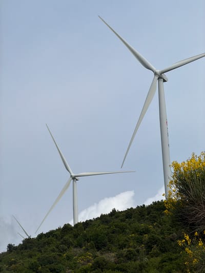 The image depicts a wind farm in Kruče - Kërruçi, Montenegro. Three white wind turbines are visible against a cloudy sky. Dense green vegetation, with some yellow flowering bushes, lines the right side of the image, partially obscuring the turbines. The angle is tilted upwards, giving a worm's-eye view of the wind turbines extending into the sky.