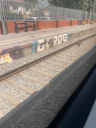 The image captures a view from a train window looking out at a train platform and tracks. The platform is lined with gravel, and a concrete barrier separates it from the tracks. Graffiti in white and blue is visible on the side of the platform barrier. Behind the platform, there is a brick wall topped with a metal fence. Two dark metal benches are situated on the platform. In the background, trees and a building are visible, suggesting an outdoor urban or suburban setting. The scene is illuminated by natural daylight. This location is identified as Cerdanyola del Vallès, Spain.