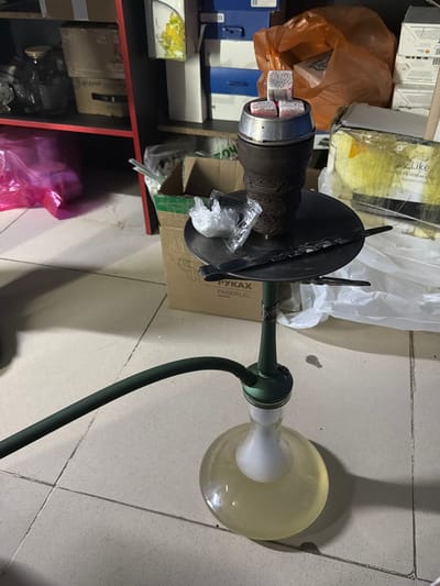 The image shows a shisha pipe setup on a tiled floor. The shisha pipe is dark brown and green, with a round glass base containing a light yellow liquid. Three white charcoal cubes are placed on top of the shisha bowl, which is a dark metallic color. A black hose extends from the pipe, running horizontally across the lower left of the frame. In the background, shelves are visible, filled with various items including boxes, a pink plastic bag, and an orange plastic bag. The overall environment appears to be indoors, possibly a storage area or a room with cluttered shelves. The lighting is dim, creating shadows on the floor.
