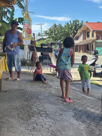 The image depicts an outdoor daytime scene in Burgos, Philippines, under a clear blue sky. Four individuals are present on a grey hexagonal-patterned pavement. On the far left, an adult male, wearing a blue tank top, faded denim shorts, and a dark baseball cap, stands smiling towards the camera, holding a clear plastic bag. A gold-toned watch is visible on his left wrist. Behind him, a tall pole displays a white banner with red text, including "OMNIFEST PANALO SALE."

In the center-left foreground, a young barefoot child in a red sleeveless top and dark blue pants is seated on the pavement, smiling. To their right, a slightly older child, wearing a light blue sleeveless top with cartoon prints and a light purple patterned skirt, stands with their back largely to the camera, holding a pink object in their left hand. Further to the right, a third barefoot child, dressed in a green t-shirt with "HOP!" and a yellow smiley face, and white shorts, stands smiling towards the center.

In the mid-ground, a black metal barbecue grill is situated behind the seated child. Beyond the pavement, a paved road is visible. Parked on or beside the road are a dark-colored tricycle, which has "BIBLE BAPTIST CHURCH FAMILY" text partially visible on its roof, and a white scooter or motorcycle. The background features a two-story building with reddish-brown brick-like siding and a prominent red corrugated metal roof, alongside lush tropical vegetation including palm trees.