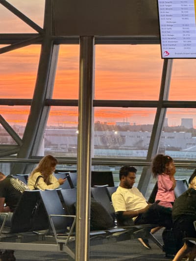 An interior shot captures a sunset viewed through a large airport window, with an overhead flight information display board partially visible. The sky is filled with vibrant orange and pink hues. In the foreground, several individuals are seated in airport waiting area chairs. A woman with long brown hair is seated in a yellow sweater, looking at her phone. To her right, a man in a white t-shirt and dark pants is seated, looking down. A young girl in a pink shirt stands to the right of the man, her hands near her face. Another person wearing a black t-shirt with a white graphic is seated to the left of the woman. The architecture of the airport, with metal framing and seating, is visible. The flight information board displays destinations such as Tenerife, Tangier, Alicante, and Amsterdam. The sign below the board indicates "Brussels Airport".