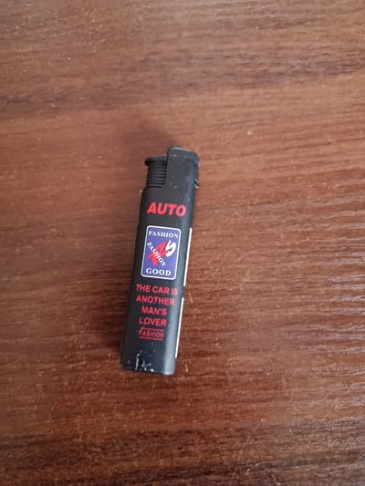 A black disposable lighter with red and blue text is lying on a wooden surface. The lighter has text printed on it, including "AUTO", "FASHION", "GOOD", and "THE CAR IS ANOTHER MAN'S LOVER". There is a graphic on the lighter that includes the word "FASHION" repeated twice, with a stylized "99" in between. The wooden surface appears to be a table or desk, with a visible wood grain pattern. The lighting suggests it is daytime. No people are visible in the image. The scene is a close-up shot of the lighter against the wooden background.