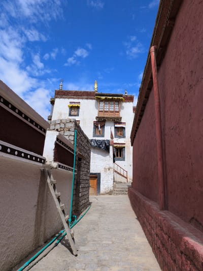 This is a photograph of an alleyway leading towards a white building with a brown tiled roof and ornate windows. The building has several stories and is decorated with flags and what appear to be prayer wheels on the roof. The alley is paved with cobblestones and is flanked by two walls. The wall on the left is white with dark brown and white decorative bands. A wooden ladder leans against this wall, alongside some turquoise pipes. The wall on the right is a deep red color and appears to be made of brick. The sky is a vibrant blue with scattered white clouds. The image is taken from a low angle, looking up towards the building and the sky. This scene is located in Choma, India. There are no people visible in the photograph.