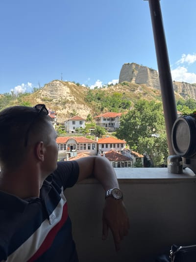A male individual is observed from a rear-right perspective, gazing towards a panoramic view. He is wearing a dark polo shirt with a vertical white stripe bordered by a red stripe on his left side, and black sunglasses are positioned on his head. A silver-toned wristwatch is visible on his left wrist, which rests on a light-colored concrete parapet. To the far right, a dark cylindrical structural pillar extends vertically. On the parapet, a small wooden decorative barrel with a metal band and a small cylindrical metallic object are present. The middle ground reveals the city of Melnik, Bulgaria, characterized by traditional houses with orange-red tiled roofs, light-colored walls, and numerous windows, interspersed among mature green trees. The background is dominated by prominent, light-colored sandstone formations, consistent with the Melnik Earth Pyramids, which are partially covered with sparse green vegetation, all set against a clear blue sky with scattered white cumulus clouds.