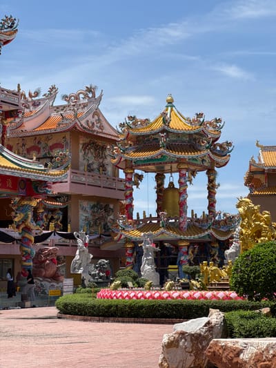 In Ang Sila, Thailand, this image captures a vibrant Chinese temple under a clear blue sky with sparse clouds. The temple's architecture is characterized by ornate, multi-tiered roofs adorned with colorful dragons and intricate carvings. A large, golden bell hangs prominently within a pavilion supported by red and white spiral columns, also decorated with dragon motifs.
In the foreground, a red brick courtyard leads to a manicured garden area featuring low green bushes and a decorative border of pink lotus-like structures. Several stone sculptures are visible, including white figures and guardian lion statues. To the right, a large golden sculpture of mythical creatures stands.
The temple's facade is richly decorated with murals depicting figures and scenes, further enhanced by dragon sculptures on pillars and along the rooflines. In the middle ground, several individuals are seen walking on steps near the temple entrance. The overall scene conveys a sense of cultural richness and spiritual significance, bathed in bright daylight.