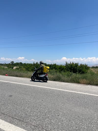 The image shows a person riding a scooter on a paved road. The rider is wearing a helmet and dark clothing, with a bright yellow backpack attached to the back of the scooter. The scooter is white and black. The road has a white line marking the lane. To the left of the road, there is a stretch of green grass and vegetation, with trees and bushes. In the background, there are more trees and hills, and a bright blue sky with some white clouds. There are also power lines visible in the upper left portion of the image. The scene appears to be in a rural or semi-rural area. The lighting suggests it is daytime, likely sunny. The image is oriented vertically, with the sky at the top and the road below. There is a small red object on the side of the road. A sign with yellow text is partially visible on the side of the road, but the text is unreadable.