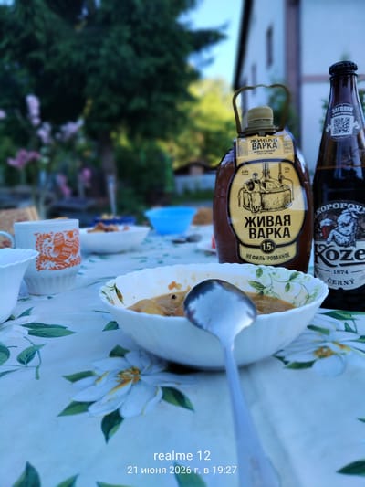 In an outdoor setting, a table is set with a meal and drinks. In the foreground, a white bowl with a floral pattern contains a portion of stew with a spoon resting inside it. To the left, a white mug with an orange bird motif sits on a tablecloth patterned with white flowers. In the background, two bottles are visible: one with a brown label and a distinctive handle, and another brown beer bottle with a label reading "Koze". The background also shows greenery and the side of a building, suggesting a residential or recreational area. The timestamp "realme 12 21 июня 2026 г. в 19:27" is superimposed on the bottom right of the image. There is no indication of the location being the city of Космос-2 Снт, Russia.