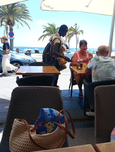 This bright, sunlit image captures an outdoor cafe scene by the sea, likely in Puerto del Carmen, Spain. In the foreground, a woven beige bag with brown straps and a blue hat are resting on a dark patterned chair.
The main focus is a dark-skinned man, wearing a patterned shirt and a backpack, approaching a table where two diners are seated. The woman, wearing a pink top and sunglasses, is laughing heartily while holding a glass with a drink and a straw. She is looking towards a man in a light blue floral shirt, seen from behind. A condiment set is on their table.
Beyond the cafe seating area, a street is visible with a blue car and a scooter. A person in a black t-shirt and white pants, carrying a white bag, walks past the car. The background reveals the serene blue ocean with scattered straw beach umbrellas and rocky formations. Tall palm trees punctuate the clear, bright blue sky, indicating a sunny day.
Notable details include a red and blue 'no stopping' sign near the street, and the word "San Miguel" (appearing as "lqpiuMnaS" as it's upside down) printed on the white parasol providing shade over the cafe. The overall atmosphere is relaxed and cheerful.