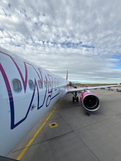 An exterior, eye-level shot shows a Wizz Air airplane parked on an airport tarmac under a partly cloudy sky. The fuselage of the plane, which is white with a large purple and pink Wizz Air logo, occupies the left and center portions of the frame. Several windows are visible along the fuselage. The right side of the plane features a bright pink engine nacelle with a silver fan. The tarmac is gray concrete with yellow and white lines. In the background, a mobile staircase is attached to the plane, and other airport equipment and a distant building are visible. The scene appears to be an airport in or near Künersberg, Germany, given the Wizz Air livery and common airport features.