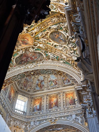 This is a close-up, upward-facing view of the ornate ceiling of the Church of San Giorgio Martire in Mozzo, Italy. The ceiling is a complex architectural and artistic composition featuring a dome with painted frescoes and intricate stucco work, all heavily adorned with gold accents.
The frescoes depict religious scenes with numerous figures in traditional attire, rendered in rich colors. These paintings are framed within oval and rectangular cartouches, surrounded by elaborate gilded stucco ornamentation, including cherubs, drapery, and architectural motifs. The stucco work itself is highly detailed, creating a sense of depth and movement.
The viewpoint suggests the photographer is standing within the nave of the church, looking up towards the crossing or a dome. A large dark wooden architectural element, possibly a beam or part of a choir loft, is visible in the foreground on the left, partially obscuring the ceiling. On the right side of the image, a large, light-colored sculpted figure, possibly a saint or angel, is positioned on a column, further emphasizing the grand scale and decorative richness of the interior. The overall impression is one of opulent religious artistry and architecture.