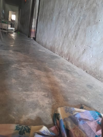 This image depicts a dimly lit concrete corridor within a building in Ifo, Nigeria, viewed from a low angle. The floor is unpolished concrete, exhibiting mottled patterns of gray and brown with some reflective areas. The walls are rough, unpainted concrete or plaster, with the right wall displaying significant texture and minor deterioration along its base. In the immediate foreground, a folded or bunched fabric, patterned with blue, purple, beige, and green floral or geometric designs, rests on the floor. Towards the midground on the left, a wooden structure, possibly a table or workbench, is visible, accompanied by several thin wooden supports leaning vertically. A small, bright green plastic cup is on the floor near the left wall, and two dark-colored sandals are positioned on the floor closer to an open doorway. Through this open, brightly illuminated doorway at the far end of the corridor, a partially obscured individual is discernible standing, with other indistinct objects visible in the background room. No direct actions or interactions between individuals are clearly depicted.