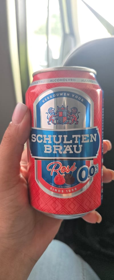 A person's hand with a light pink manicure holds a red aluminum can of "Schulten Brau" non-alcoholic rose beer. The can features a silver and blue label with two lions flanking a shield, and the text "GEBROUWEN VOOR," "SCHULTEN BRAU," "Rosé," and "0.0% ALCOHOLVRIJ." Below the "Rosé" text, it says "MET SMAAK VAN ROOD FRUIT" and "SINDS 1988." The background is out of focus, showing a light-colored curtain or blind on the left and a dark, possibly car interior, on the right. The lighting suggests an indoor environment, possibly a vehicle. There are no discernible landmarks or specific locations within Zwartewaal, Netherlands, visible in the image.
