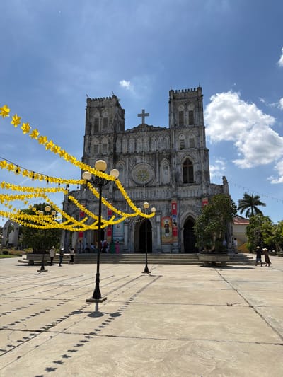 The image depicts a wide-angle view of the Church of Hội Tín in Hội Tín, Vietnam, under a bright, partially cloudy blue sky. The church, a grand stone structure with two prominent towers and a cross at its apex, dominates the background. Yellow star-shaped lanterns are strung across the foreground, creating a festive atmosphere. Several streetlights with spherical lamps are interspersed among the lanterns. The courtyard in front of the church is paved with concrete and features patterned shadows from the surrounding structures and lighting. A few people are visible in the courtyard and on the steps leading to the church entrance. To the right of the church, palm trees and other lush greenery are present. The overall scene suggests a sunny day, possibly during a celebration or event, given the decorations.