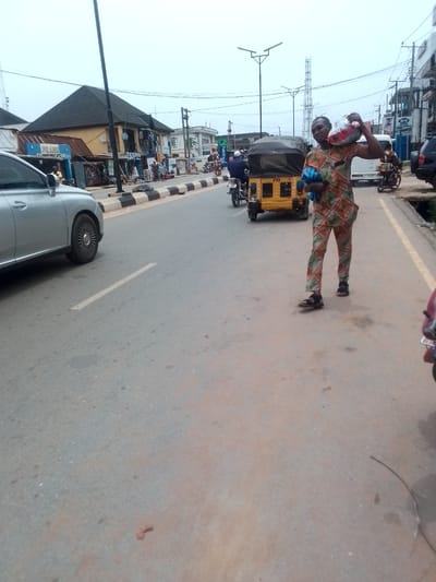 A street in Orlu, Nigeria, features a man carrying a large bag on his shoulder. He is wearing a patterned orange and green outfit with sandals. Beside him on the road is a yellow tricycle. Other vehicles, including cars and motorcycles, are on the street, and pedestrians are visible in the background near shops and buildings. Streetlights and utility poles line the road.