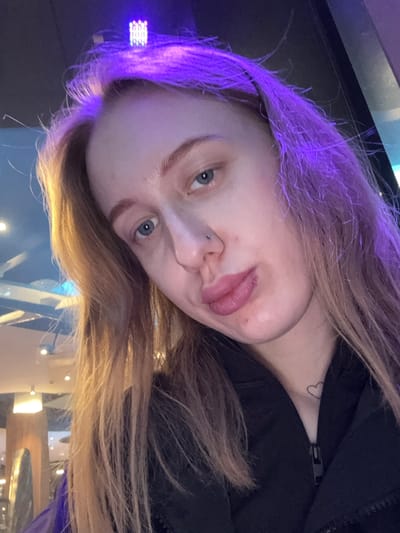 A fair-skinned individual with dyed blonde hair is facing the camera with their tongue extended. They have a pierced nose and blue eyes. A black zipper jacket is visible. A small heart tattoo can be seen on their neck. The background appears to be a brightly lit, possibly public space with blurred elements. The image is taken at an angle. There is no indication this image was taken in Ol'ginskaya, Russia.