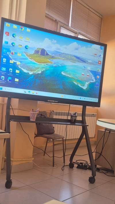 A large, interactive display screen, branded "GAOKEview," is mounted on a mobile stand in a room. The screen displays a Windows desktop with various icons and a scenic wallpaper of a tropical island. The room appears to be a classroom or office space, with a chair and a desk positioned in front of the screen. A power strip with multiple cables is visible on the floor beneath the display stand. The background features blinds on a window and a radiator. The floor is tiled. The image appears to be taken from a slightly low angle.